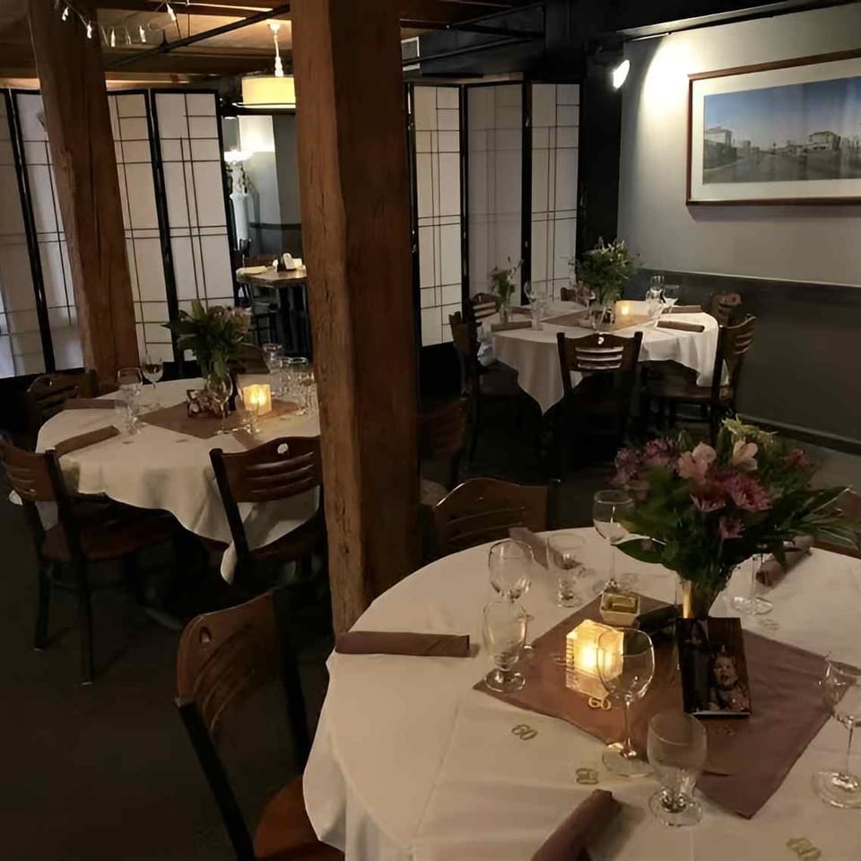 Dimly lit restaurant dining room with white-clothed tables, wooden chairs, and floral centerpieces.