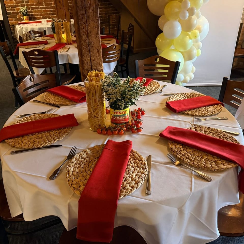 Round table set with white tablecloths, red napkins, straw placemats, and pasta-themed centerpieces.