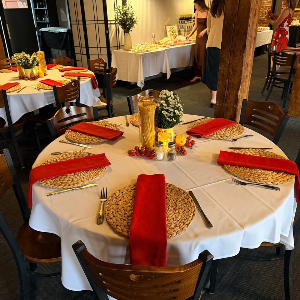 Restaurant dining room with round tables set with white tablecloths, red napkins, and wicker placemats.