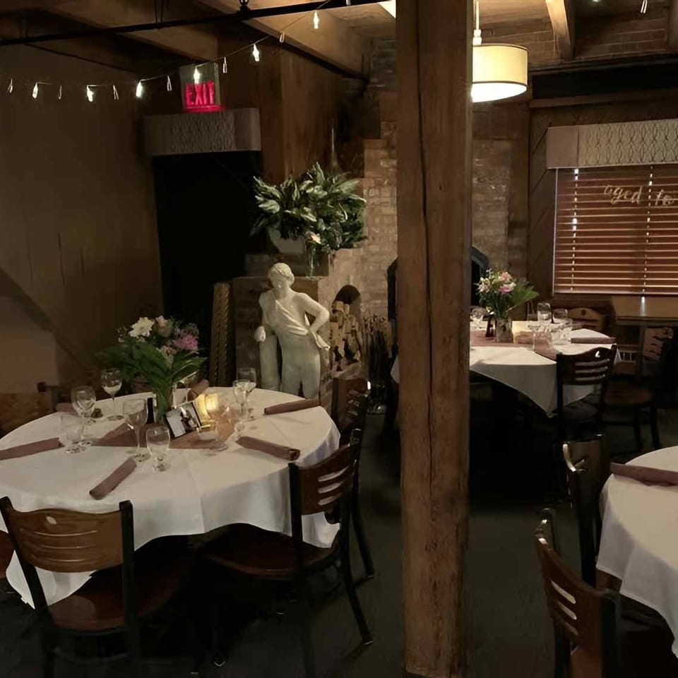 Cozy restaurant interior with white-clothed tables, wooden beams, and a classical statue under warm lighting.