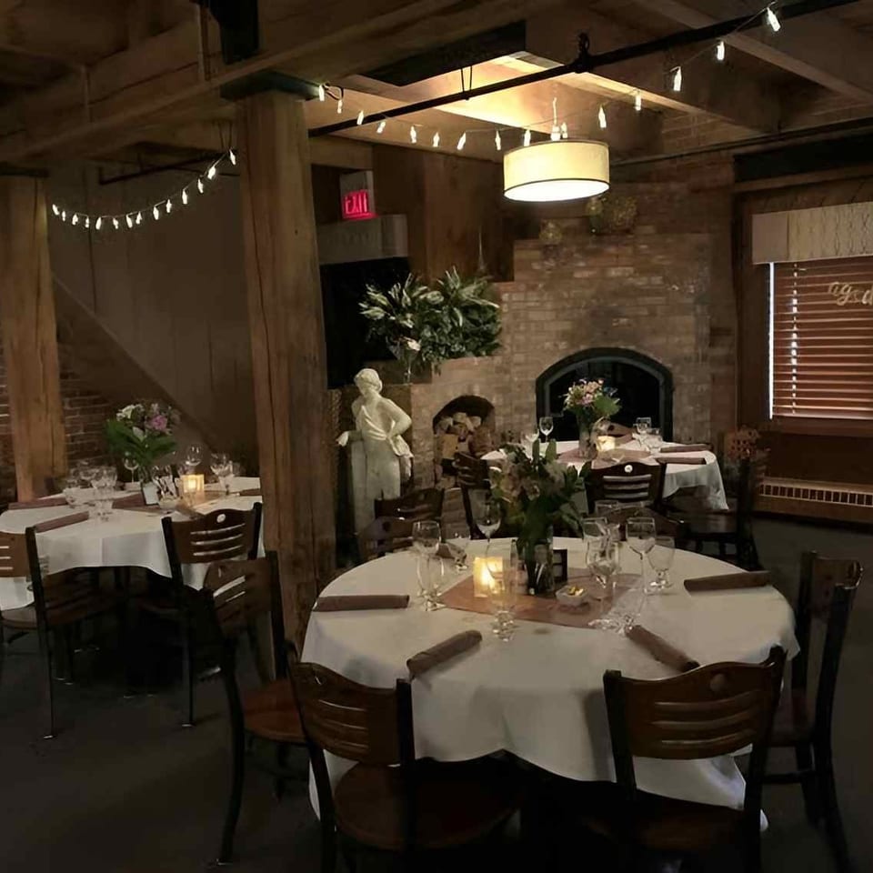 Cozy restaurant dining room with white-clothed tables, wooden beams, brick fireplace, and decorative statues.